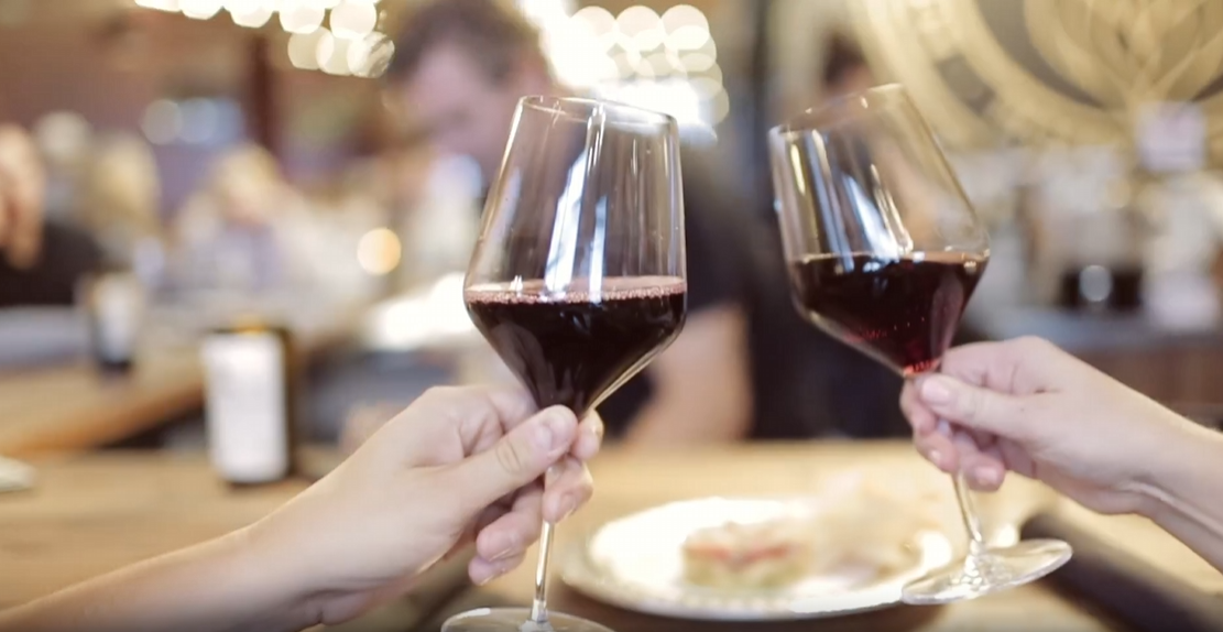 Two guests toasting glasses of red wine at the bar under warm string lights