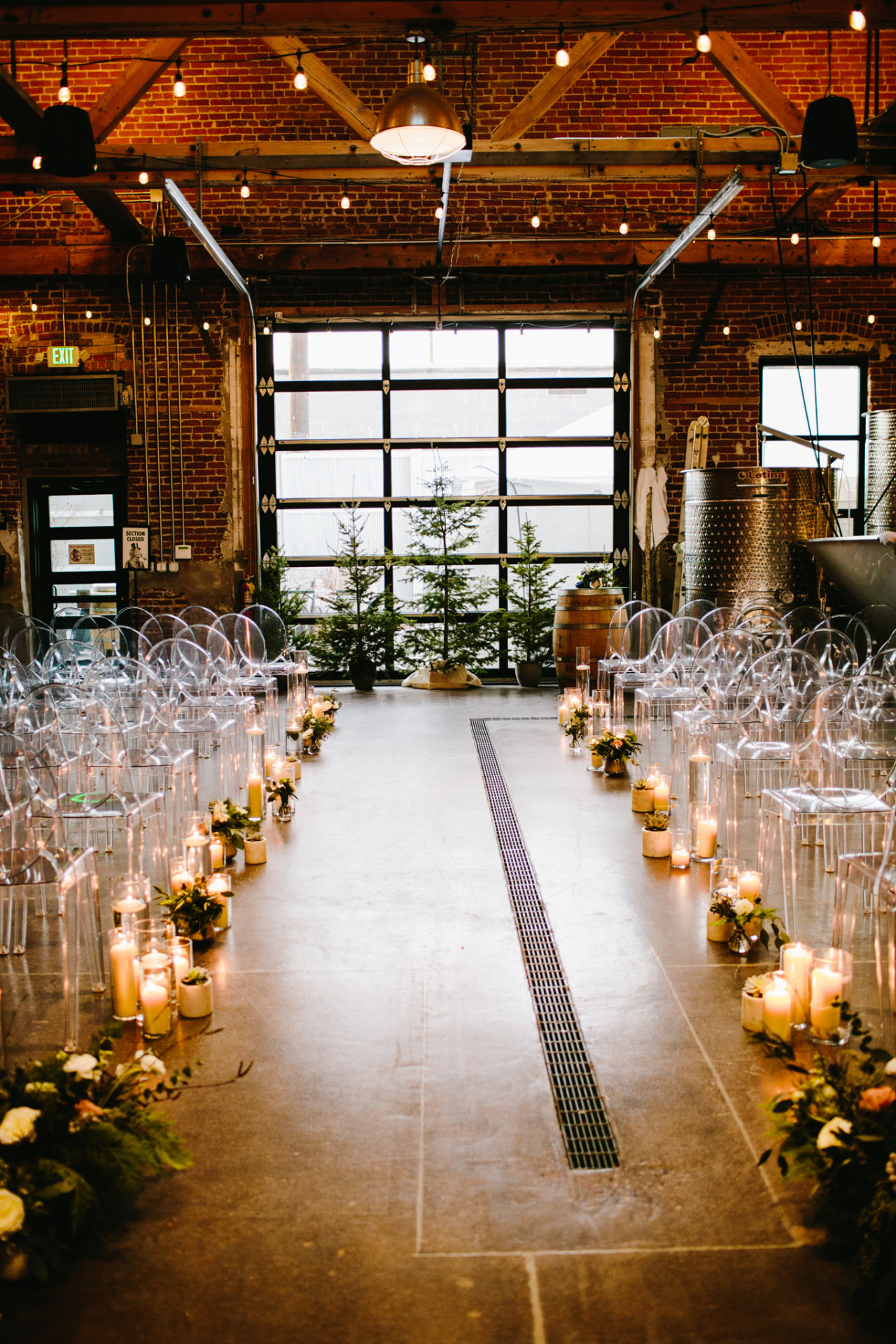Candlelit ceremony aisle lined with clear chairs and evergreens in the brick hall