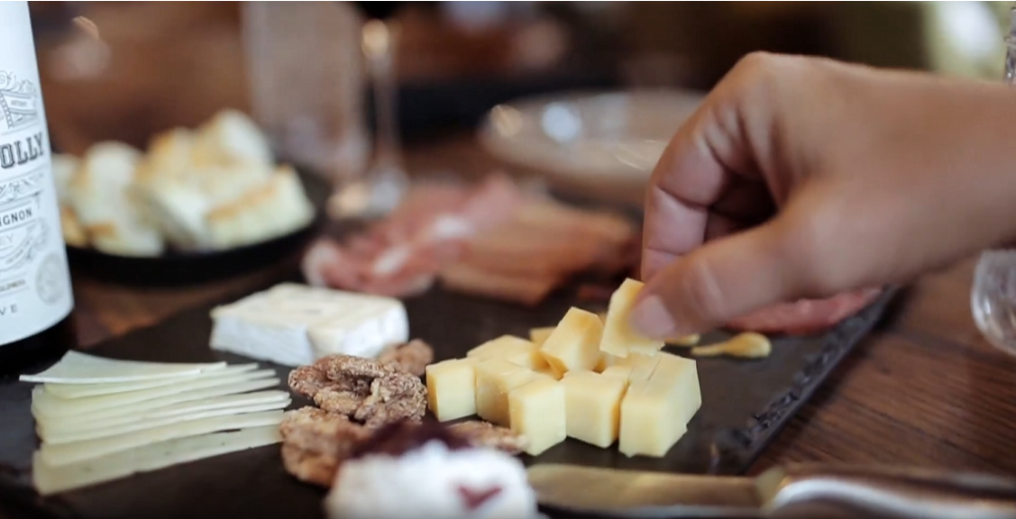 A guest reaching for cheese on a slate board with charcuterie, candied nuts and a bottle of Bigsby's Folly wine