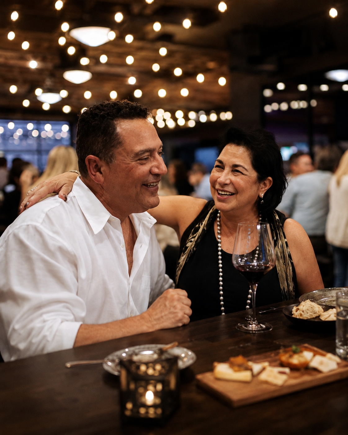 A couple laughing over a glass of red wine and a cheese board beneath warm string lights in the winery's dining room